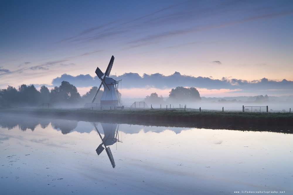 white Dutch windmill by river in misty sunrise